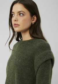 Pull en maille verte avec une finition texturée, un col rond et des manches roulées, mettant en valeur un tissu doux et confortable.