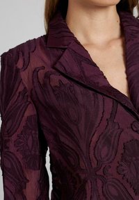 Paarse geborduurde blazer met een gestructureerd bloemenpatroon en een klassieke kraag. Lichte, doorzichtige stof met zichtbare stikdetails.