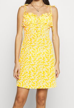 Robe fleurie jaune avec bretelles ajustables, corsage ajusté et décolleté froncé. Présente des motifs de fleurs blanches et un design à mi-cuisses.