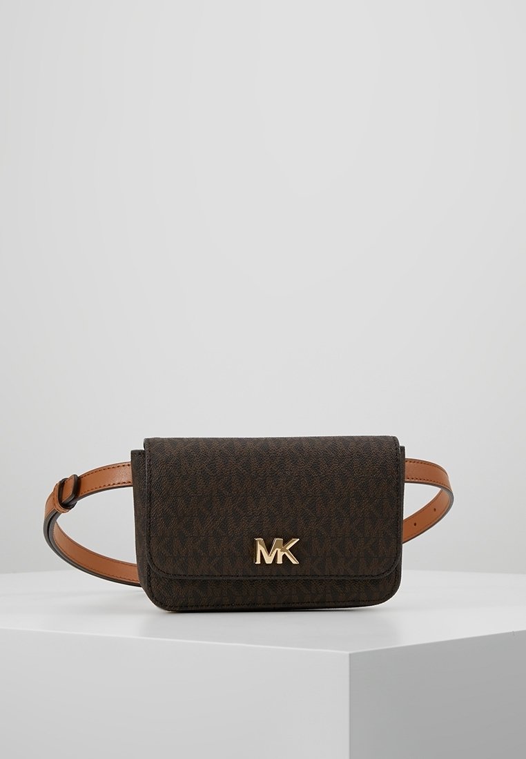 Musta ruskea Michael Kors vyölaukku, jossa on kultainen MK-logo ja säädettävä vaaleanruskea nahkahihna, valkoisella jalustalla harmaata taustaa vasten.
