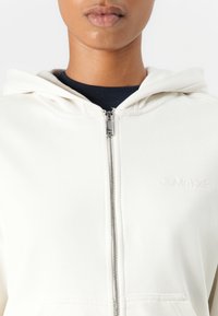 Felpa bianca con zip e cappuccio, caratterizzata da una texture liscia, dettagli cuciti e un piccolo logo inciso sul petto.