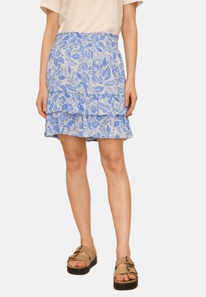 Femme portant un haut beige, une mini-jupe à volants florale bleue et blanche, et des sandales à plateforme beiges avec deux boucles.