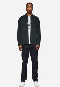 Schwarze Cordjacke mit Knopfleiste, einseitiger Brusttasche, über einem weißen Grafik-T-Shirt getragen, kombiniert mit dunklen Jeans und weißen Sneakers.