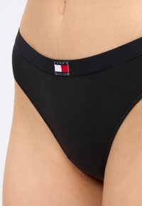 Černé kalhotky s vysokým střihem s logem Tommy Hilfiger na pružném pásku, nošené v pase na holé kůži.