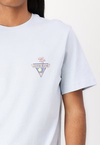T-shirt en coton bleu clair avec un graphique triangulaire représentant des palmier, un soleil et des vagues de l'océan. Texte "GUESS JEANS" dans une police contrastante.