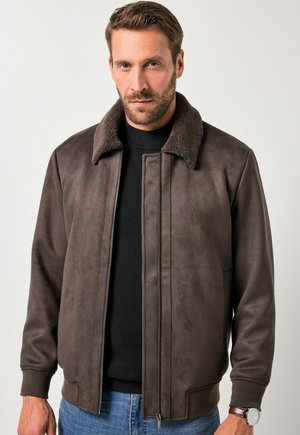 Chaqueta bomber de ante marrón con un cuello de piel oscura texturizada, cierre de cremallera en el frente y puños acanalados, combinada con un suéter negro debajo.