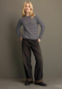 Schwarz-weiß gestreiftes Langarmoberteil mit Rundhalsausschnitt, kombiniert mit dunkelblauen weiten Jeans und schwarzen Ankestiefeln. Einheitlicher Hintergrund.