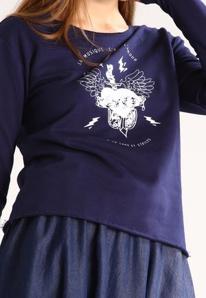 Femme portant un t-shirt bleu marine à manches longues avec un graphique de cœur ailé blanc, des éclairs et du texte en français, associé à une jupe en denim.