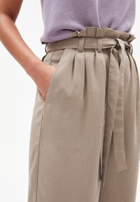 Světle béžové kalhoty s vysokým pasem, nažehleným předním dílem a látkovým opaskem. Mají boční kapsy a hladkou, jemnou texturu.