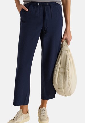 Persona che indossa pantaloni ampi blu navy texturizzati e sneaker beige, tenendo una grande borsa imbottita beige per la maniglia.