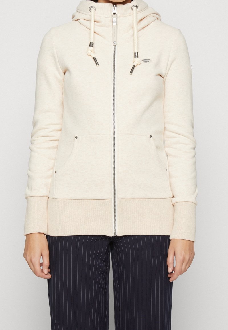 Veste à capuche zippée beige en tissu doux, dotée d'une capuche à cordon de serrage, de poches avant et d'accents métalliques. Équipée de poignets et d'un ourlet en bord-côte.