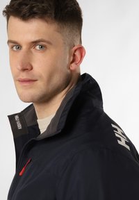 Chaqueta impermeable azul marino con cuello alto, tirador de cremallera rojo y detalles grises en el cuello. Presenta un logotipo en el hombro.