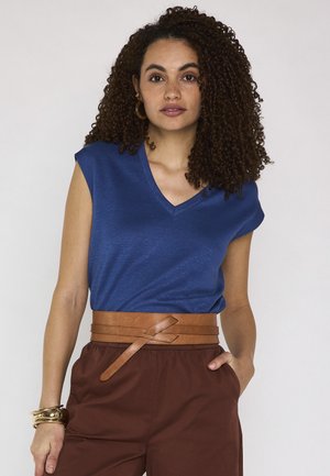 Woman with curly hair wearing a blue sleeveless V-neck top, brown high-waisted pants, wide tan belt, and gold bracelets, standing with one hand in pocket.