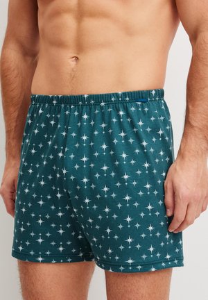 Uomo che indossa boxer verde petrolio scuro con motivi di stelle bianche, mostra il torso dalla vita a metà coscia su sfondo semplice.