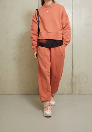 Osoba nosząca koralową bluzę Nike i dopasowane spodnie dresowe, różowo-białe sneakersy, czarną torbę na ramię, stojąca na tle jednolitej ściany.