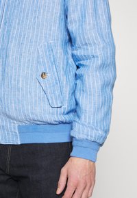 Ljust blå, randig jacka med en framficka och knappdetalj. Har ribbade ärmslut och nederkant. Tillverkad av lättviktsmaterial.