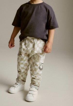 Tout-petit portant une chemise à manches courtes gris foncé, un pantalon à carreaux beige et blanc avec un patch smiley, et des baskets blanches à scratch, debout.