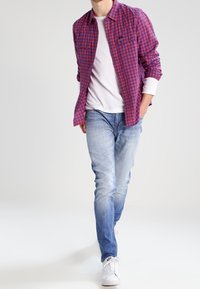 Mann, der hellblaue Jeans, weiße Turnschuhe, weißes T-Shirt und ein aufgeknöpftes rot-blau kariertes Hemd trägt, mit den Händen in den Taschen.