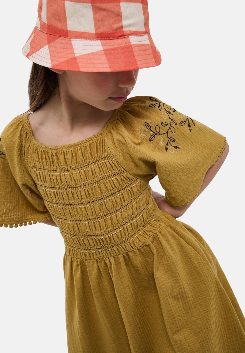 Robe jaune moutarde avec corsage smocké et manches volantées, ornée d'une broderie florale noire et d'une jupe à volants. Associée à un chapeau seau à carreaux rouges et blancs.