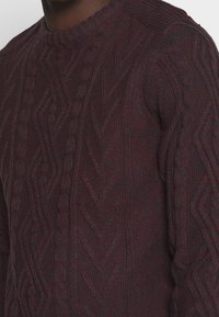 Dunkelburgunderfarbener Kabelstrickpullover mit strukturierten Mustern und geripptem Kragen. Das Material wirkt dick und hat eine gemütliche Ausstrahlung.