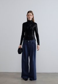 Μαύρο τοπ με παγιέτες και ψηλό λαιμό, συνδυασμένο με φαρδιά σκούρου μπλε τζιν παντελόνια. Το μοντέλο κρατάει μαύρο φάκελο. Μινιμαλιστικό φόντο.