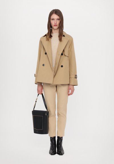 Beige dupla gombos trench kabát fekete gombokkal, krém színű pulóver, bézs nadrág és fekete vödör táska arany kiegészítőkkel; fekete magassarkú csizma.