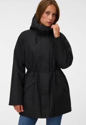 Femme portant un manteau noir à capuche avec des cordons à la taille et deux poches avant à rabat, posant avec une main près du menton.