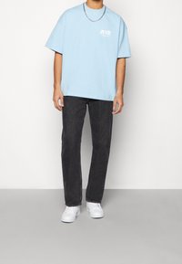 Ljusblå bomullströja med kort ärm och logotryck; kombinerad med mörka denimjeans och vita sneakers. Silverfärgad kedja.