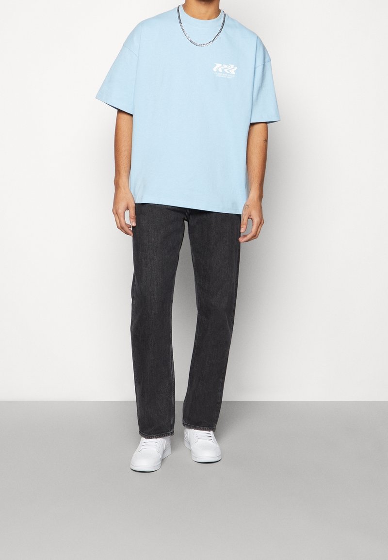 Ljusblå bomullströja med kort ärm och logotryck; kombinerad med mörka denimjeans och vita sneakers. Silverfärgad kedja.