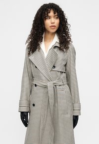 Καμπαρντίνα σε ασπρόμαυρο καρό σχέδιο, με διπλό πέτο, φαρδιές πέτσες, ζώνη και εφαρμοστή γραμμή.