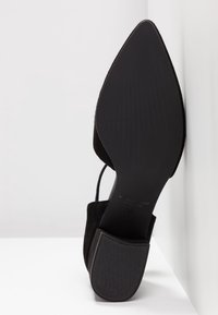 Semelle de chaussure noire à bout pointu avec un petit talon bloc, positionnée verticalement contre une surface blanche.
