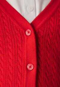 Crveni pleteni kardigan s uzorcima pletenica, dva crvena gumba zakopčana preko bijelog ovratnika košulje s gumbima.