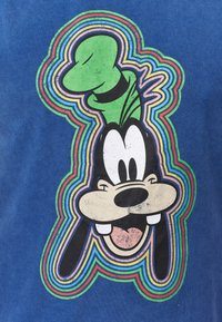 Blauwe t-shirt met een afbeelding van een lachend cartoonfiguur met grote witte ogen, een opvallende groene hoed en regenboogachtige concentrische omlijningen.