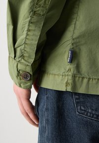 Zelená lehká bunda s detaily na manžetách, hladkým povrchem a malou značkovou etiketou. Nošena přes tmavé džíny.