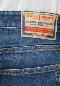 Mėlyni džinsai su kontrastinga oranžine siūle. Rudas odinis ženklas rodo "Diesel Industry" logotipą ir produkto detales.