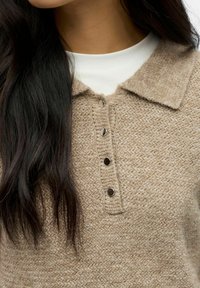 Donna con lunghi capelli scuri che indossa un maglione beige lavorato a maglia con bottoni sopra una maglietta bianca, mostrando il colletto e i bottoni.