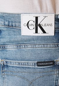 Světle modré džíny vyrobené z denimu, které mají bílou koženou etiketu s černým nápisem "CK CALVIN KLEIN JEANS" na pásku a malou značkovou nášivku na zadní kapse.