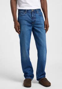 Jeans de mezclilla azul con un diseño de pierna recta, costuras amarillas en contraste y bolsillos delanteros. Llevados con zapatos marrones.