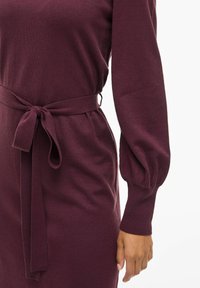 Robe en tricot bordeaux avec des manches longues, une taille ceinturée et des manches bouffantes. Texture douce et silhouette ajustée mettant en valeur la taille.
