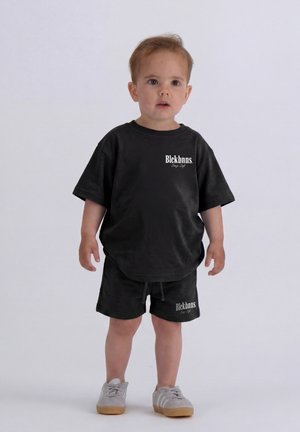 Peuter die rechtop staat, draagt een zwarte oversized T-shirt en korte broek met witte tekst, en lichtgrijze sneakers met rubberen zolen tegen een egale achtergrond.
