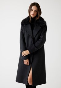 Caban lung din lână neagră cu guler din blană sintetică, design croit, fante laterale vizibile și rever structurat, evidențiind o textură netedă și elegantă.