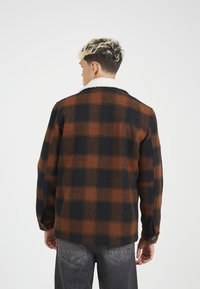 Geruite fleecejack met een ontwerp in bruin, zwart en oranje, voorzien van een pluizige witte kraag en knoopdetails op de mouwen.