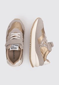Sneaker beige e oro con tomaia in suede, dettagli metallici, una fascia in velcro e una suola texturizzata con leggera gomma.