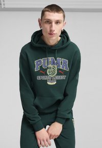 Felpa con cappuccio verde scuro con branding PUMA, presenta una tasca frontale a marsupio, coulisse e polsi a coste. Tessuto morbido e texturizzato.