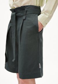 Tmavě zelené lněné šortky s širokým pasem, samostatným opaskem na zavázání a bočními kapsami. Mají strukturovanou tkaninu a decentní značkovou etiketu.