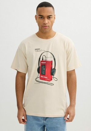 Joven con camiseta beige con gráfico de reproductor de cassette rojo y auriculares negros, de pie frente a un fondo blanco liso.