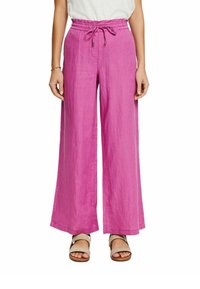 Femme portant un pantalon en lin rose taille haute à jambes larges avec taille à cordon et sandales beiges, debout devant un fond blanc.