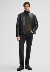 Schwarze Lederjacke mit hohem Kragen über einem dunkelgrünen gerippten Pullover, kombiniert mit schwarzen Jeans und schwarzen Schnürstiefeln.