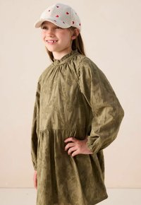 Lächelndes Mädchen in einem hellgrünen, blumigen Kleid mit langen Ärmeln und einer weißen Mütze mit kleinen roten Herzen, steht mit der Hand auf der Hüfte.