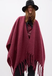 Bordo pončo od mekog, teksturiranog materijala s dugim resicama na rubu, nosi se preko tamne košulje s kragnom. Crni šešir stvara kontrast.